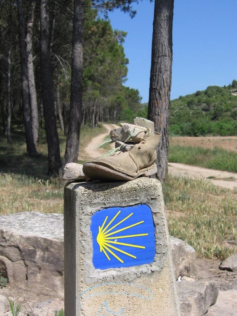 Santiago de Compostelan vaelluksen sininen reittimerkki, jossa keltainen simpukkasymboli. Kivisen reittimerkin päällä on vanha vaelluskenkä.